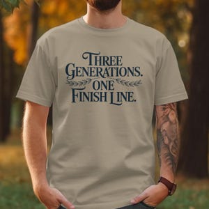 Peut inclure: T-shirt beige avec l'inscription bleu marine "THREE GENERATIONS. ONE FINISH LINE.". Le texte est disposé sur trois lignes avec des ornements de feuilles. Le t-shirt est à manches courtes.