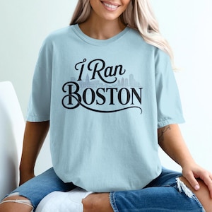 Può includere: T-shirt azzurro con la scritta "I Ran Boston" in corsivo nero e una grafica dello skyline di Boston. La maglietta ha maniche corte e una vestibilità ampia.