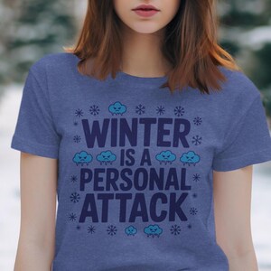 Może przedstawiać: Niebieska koszulka z napisem "WINTER IS A PERSONAL ATTACK" w kolorze białym i niebieskim. Projekt zawiera płatki śniegu i grafiki wściekłych chmur. Koszulka ma okrągły dekolt i krótkie rękawy.