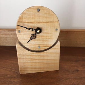 Pode incluir: Um relógio de madeira castanho-claro com um mostrador redondo e uma base retangular. Os ponteiros do relógio são pretos e o centro é de cor latão. O design é simples e o relógio tem um aspeto rústico.