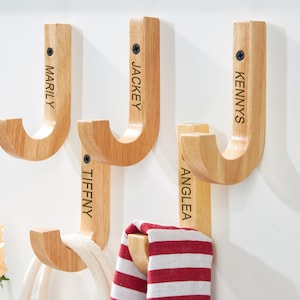 Puede incluir: Ganchos de pared de madera en forma de J, cada uno con un nombre grabado en negro: Marily, Jackey, Kennys, Tiffny y Anglea. Algunos ganchos sostienen una cuerda blanca y una bufanda a rayas rojas y blancas. Una rosa melocotón está en la esquina inferior izquierda.