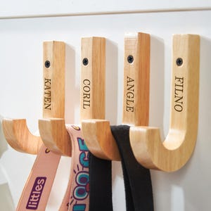 May include: Four wooden wall hooks with engraved names: Katen, Coril, Angle, and Filno. Each hook is a light wood color with a curved design. Various straps and belts hang from the hooks, including a pink strap with the word "littles".