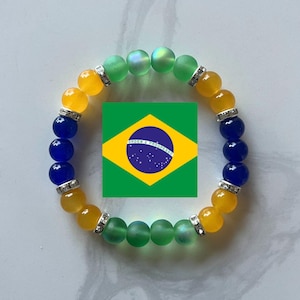 Könnte beinhalten: Ein Armband mit gelben, blauen und grünen Perlen, getrennt durch silberfarbene Abstandshalter. Das Armband ist um eine brasilianische Flaggen-Grafik angeordnet, wobei die Farben das Design der Flagge widerspiegeln. Das Armband ist wahrscheinlich für patriotisches Tragen gedacht.