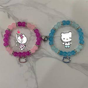 May include: Two beaded bracelets, one pink and purple, the other blue and turquoise, connected by a heart-shaped clasp. Each bracelet features a charm and a cartoon character. The bracelets are on a white marble surface.