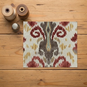 Puede incluir: Una muestra de tela cuadrada con un patrón ikat en tonos rojo intenso, marrón oscuro y amarillo apagado sobre un fondo blanco roto. Dos carretes de hilo de madera y un pequeño dedal de metal están colocados cerca de la esquina superior izquierda.