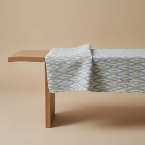 May include: A light-colored wooden bench with a flat top and a single vertical support leg, draped with a woven fabric runner featuring a repeating diamond pattern in shades of light blue, white, and muted gold across the entire surface.