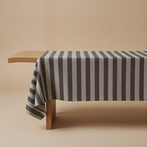 Puede incluir: Un mantel rectangular con un patrón repetitivo de rayas verticales en gris carbón oscuro y gris claro. La tela está colocada sobre un banco de madera, mostrando un tejido texturizado que cubre la parte superior y un lado del mueble.