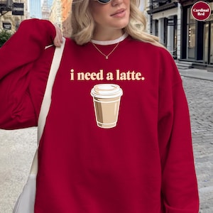 May include: A crimson sweatshirt featuring the text "i need a latte." above a graphic of a takeaway coffee cup. The cup is beige with a white lid. The sweatshirt has a white collar and is a solid colour.