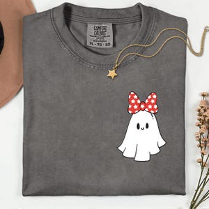Peut inclure: Un t-shirt gris Comfort Colors avec un motif de fantôme blanc portant un nœud rouge à étoiles blanches. Un collier pendentif étoile dorée est posé sur le t-shirt. Le t-shirt est de taille XL-TG-EG.