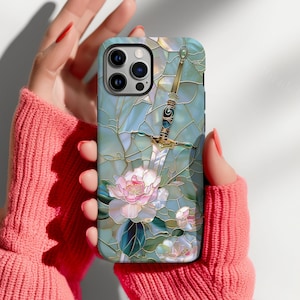 May include: A phone case featuring a stained-glass design with a sword and pink flowers. The case has a light blue background with gold accents. The phone case is held by a person wearing a pink sweater.