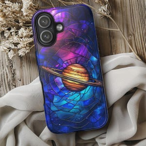 May include: A phone case featuring a stained-glass design of a planet with rings, set against a backdrop of a nebula in shades of purple, blue, and orange. The case has a glossy finish and a dark blue border.