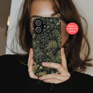 May include: A black phone case with a floral pattern featuring green and yellow flowers and foliage. The case is held up in front of a person's face. The text on the case says "Snail Paradise Co."
