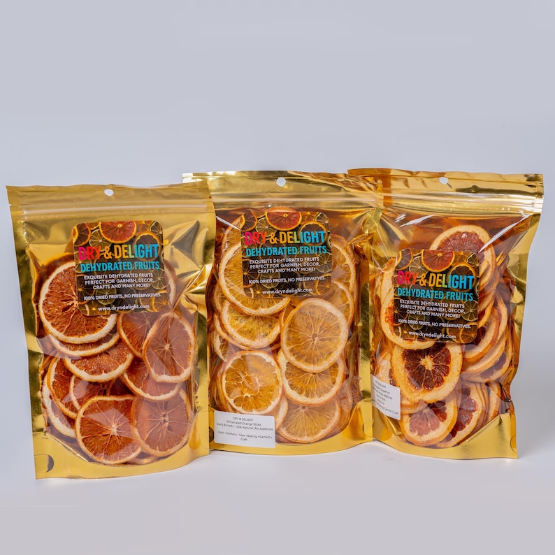 Op de afbeelding: Drie gouden foliezakken gevuld met gedroogde sinaasappelschijfjes. Elke zak heeft een label met de tekst "Dry & Delight Dehydrated Fruits". De sinaasappelschijfjes zijn in verschillende tinten oranje en bruin.