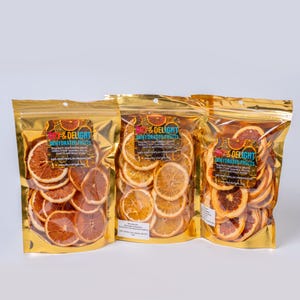 Op de afbeelding: Drie goudfolie zakken gevuld met gedroogde sinaasappel- en grapefruit schijfjes. Elke zak heeft een label met de tekst "So & Delight Dehydrated Fruits". De gedroogde fruitschijfjes zijn in nette rijen gerangschikt.