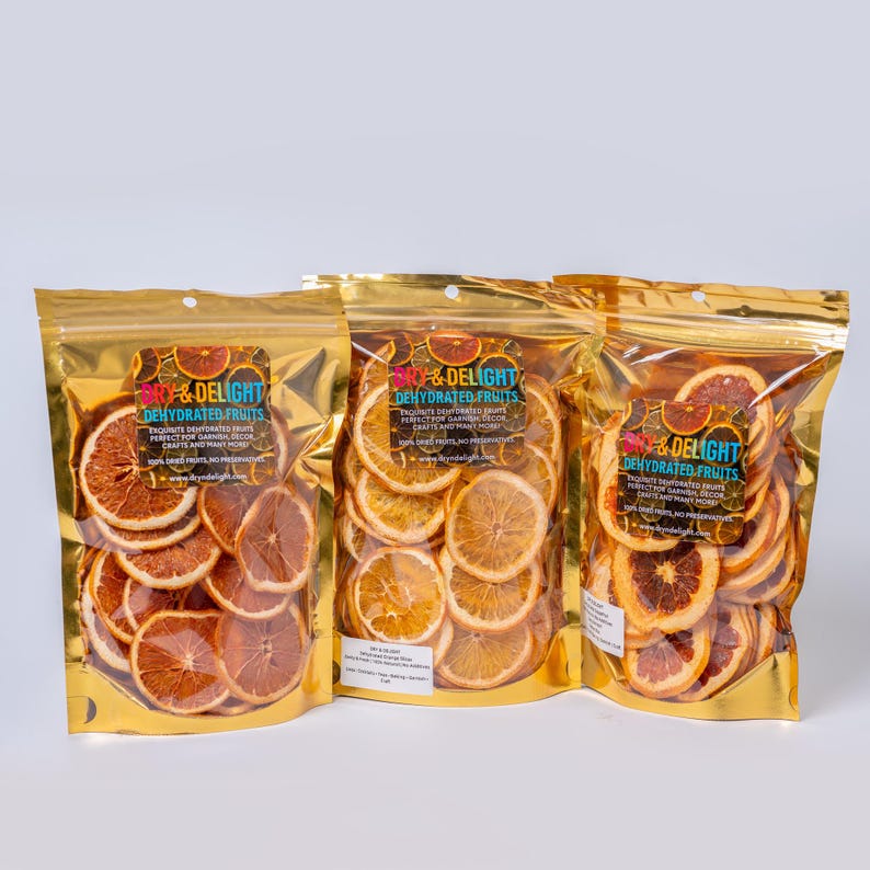 Op de afbeelding: Drie goudfolie zakken gevuld met gedroogde sinaasappel- en grapefruit schijfjes. Elke zak heeft een label met de tekst "So & Delight Dehydrated Fruits". De gedroogde fruitschijfjes zijn in nette rijen gerangschikt.