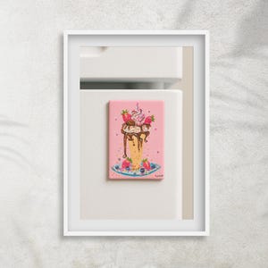 May include: A rectangular magnet featuring a colorful illustration of a strawberry sundae. The sundae has whipped cream, chocolate sauce, and fresh strawberries. The magnet is on a pink background and is framed in white.