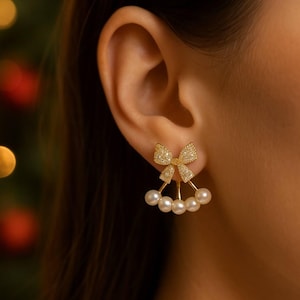 May include: Gold bow earrings with pearl clusters. The bow is set with small, sparkling stones. The earrings are designed to be worn on the earlobe, with pearls hanging below. A festive and elegant jewellery piece.