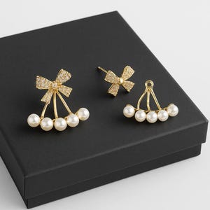May include: Gold-coloured bow earrings with faux pearls. The earrings feature a bow design at the top, embellished with small, sparkling stones. Below the bow, three or four faux pearls hang from delicate gold-coloured wires.