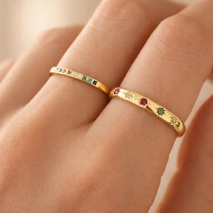 Può includere: Due anelli d'oro. Un anello presenta una fila di piccole pietre preziose colorate. L'altro anello ha pietre preziose e disegni a stella. Le pietre sono rosse, verdi e trasparenti. Gli anelli sono su un dito.