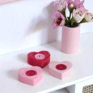May include: A display of heart-shaped candles in shades of pink and red, with a small pink vase holding pink flowers. The candles have a small candle inside. The items are arranged on a white surface.