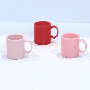 May include: Three small, solid-colored mugs. Two are a light pink, and one is red. Each mug has a rounded handle. The mugs are arranged on a white surface against a light blue background.