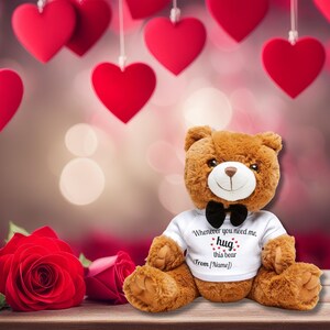 Op de afbeelding: Een bruine teddybeer met een wit t-shirt met de tekst "Whenever you need me, hug this bear (From [Name])". De beer heeft een zwarte vlinderdas en is omringd door rode harten en rozen.