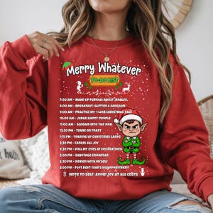 Op de afbeelding: Rode sweatshirt met de tekst "Merry Whatever" en een humoristische to-do lijst voor de feestdagen. De lijst bevat activiteiten als "Word boos wakker over jingles" en "Annuleer alle vreugde."
