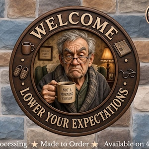 Op de afbeelding: Een rond houten wandbord met de tekst WELCOME en LOWER YOUR EXPECTATIONS. Het midden toont een ouder persoon met een bril die een mok vasthoudt met de tekst NOT A MORNING PERSON, omringd door iconen van een mok, pantoffels, krant en bril.