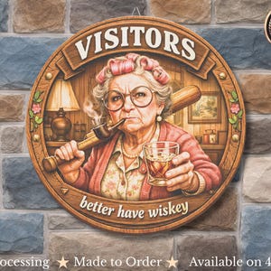 Op de afbeelding: Een rond houten bord met de tekst "VISITORS" en "better have wiskey". De afbeelding toont een oudere vrouw met een honkbalknuppel en een glas whisky. Het bord hangt aan een stenen muur.