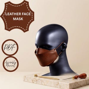 Pode incluir: Uma máscara facial de couro castanho com detalhes perfurados numa cabeça de manequim preta. A imagem inclui etiquetas de texto LEATHER FACE MASK, PDF e Assembly Manual, juntamente com uma ferramenta de metal e um furador de madeira.