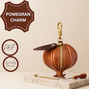 Peut inclure: Un charm en cuir marron en forme de grenade avec une fermeture éclair en laiton, une feuille en cuir marron foncé et un mousqueton en laiton. Le texte POMEGRAN CHARM, PDF et Assembly Manual est visible dans le coin supérieur gauche.