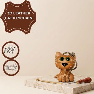 May include: A tan leather cat keychain with large green eyes, a black nose, and visible white stitching. The image includes text reading 3D LEATHER CAT KEYCHAIN, PDF, and Assembly Manual, along with a metal tool and a wooden-handled awl.