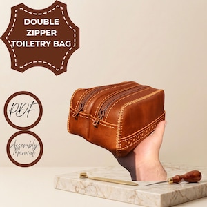 May include: A brown leather double zipper toiletry bag featuring visible stitching along the edges and sides. The image includes text labels for DOUBLE ZIPPER TOILETRE BAG, PDF, and Assembly Manual, alongside a metal tool and a wooden awl.