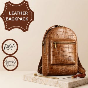 Puede incluir: Una mochila de cuero marrón con textura de piel de cocodrilo, con un bolsillo frontal con cremallera y herrajes en tono latón. La imagen incluye etiquetas de texto LEATHER BACKPACK, PDF y Assembly Manual, junto con una herramienta de metal.