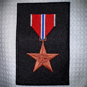 May include: A black patch featuring a bronze star pendant suspended from a red, white, and blue ribbon. The star has a detailed center design. The patch is rectangular with a stitched border.