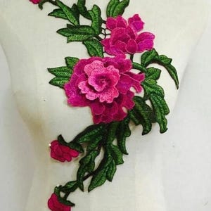 May include: Embroidered floral applique featuring vibrant fuchsia flowers and green leaves. The design includes a large central bloom and smaller buds, with detailed stitching creating texture. This decorative element is suitable for fabric embellishment.