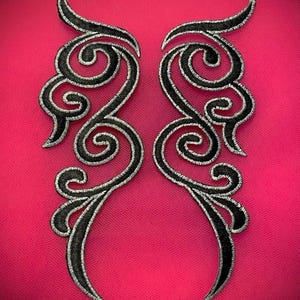 May include: Two symmetrical black embroidered appliques with a silver outline, featuring a swirling, ornate design. The appliques are set against a vibrant pink background, creating a striking contrast. The design is reminiscent of vintage or gothic styles.