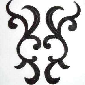 May include: Two matching, dark brown, decorative metal scrollwork pieces. Each piece features a series of curved lines and swirls, creating an elegant, symmetrical design. The pieces are set against a plain white background.