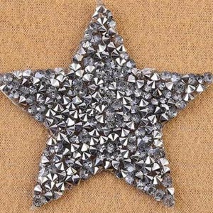 May include: A silver star-shaped embellishment covered in small, reflective rhinestones. The star is set against a textured, tan background. This decorative element could be used for crafts, fashion, or holiday decor.