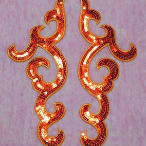 May include: Two ornate, symmetrical appliques with a swirling design. The appliques are covered in orange sequins and outlined with gold trim. The background is a light purple color.