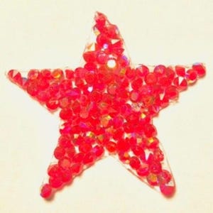 May include: A red star-shaped embellishment covered in small, sparkling red rhinestones. The star has a clear, slightly raised border, and the rhinestones are densely packed, creating a glittering effect. The background is a soft, off-white color.
