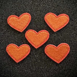 May include: Five orange heart-shaped patches with a textured design and a red border are arranged on a dark gray surface. These embroidered patches are suitable for fabric decoration and craft projects.