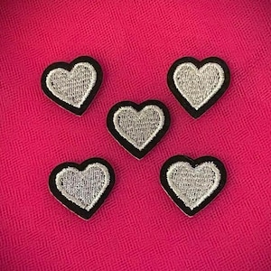 Può includere: Cinque toppe a forma di cuore con ricamo argentato e bordi neri sono disposte su uno sfondo rosa strutturato. Le toppe sono distanziate in modo uniforme, mostrando i dettagli ricamati.