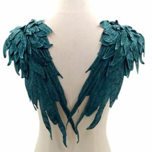 May include: Teal-colored decorative angel wings. The wings are made of a textured fabric, with a layered feather design. The wings are attached to a mannequin form, showcasing the shoulder and back placement.