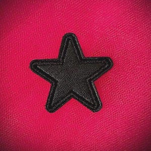 May include: A black star-shaped patch with a textured surface and stitched border is set against a vibrant pink background. The star is a solid color, and the background has a subtle, textured pattern.