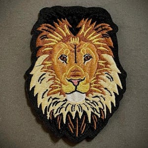 May include: Embroidered patch featuring a lion's head. The lion has a tan face, a brown mane, and a black outline. The patch is designed with detailed stitching, showcasing the lion's features.