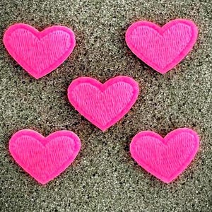 May include: Five bright pink heart-shaped patches with a textured, embroidered appearance. The hearts are arranged against a speckled, dark gray background, creating a contrast that highlights their vibrant color and shape.