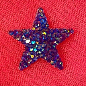 May include: A blue star-shaped ornament covered in iridescent rhinestones. The star is set against a red, textured background. The rhinestones reflect various colors, creating a sparkling effect. This decorative item could be used as a brooch or embellishment.