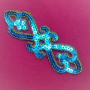 May include: A blue and white sequined applique with a swirling, symmetrical design. The applique features a dark blue outline with gold trim and is set against a pink background. The design includes a central star-shaped element.