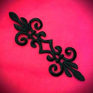 Puede incluir: Adorno negro simétrico con extremos enrollados y un diseño central en forma de diamante, sobre un fondo rosa brillante. Este elemento decorativo es probablemente para manualidades o embellecimiento.
